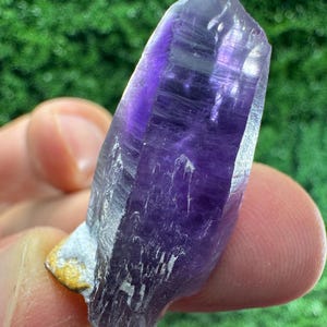 Può includere: Una punta di cristallo di ametista viola traslucido con una forma esagonale naturale e inclusioni interne visibili. Il cristallo presenta una tonalità viola intenso che sfuma verso una punta più chiara, con una piccola macchia di matrice color cuoio alla base.