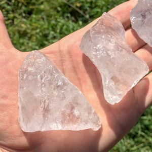 May include: Three clear quartz crystal chunks, with rough edges and a translucent appearance, are displayed on a hand.
