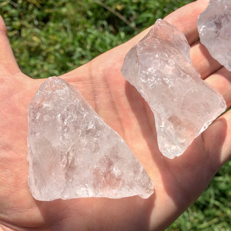 May include: Three clear quartz crystal chunks, with rough edges and a translucent appearance, are displayed on a hand.
