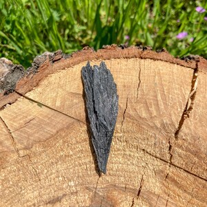 Pu&ograve; includere: Una roccia di colore grigio scuro, di formazione naturale, simile a un cristallo, con una superficie ruvida e testurizzata. La roccia &egrave; appoggiata su una superficie di legno marrone chiaro.