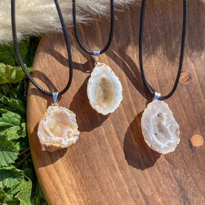 May include: Three agate crystal pendants on black cords. Each pendant is a natural, raw, and unique piece of agate with a white and brown colour scheme. The pendants are displayed on a wooden surface.