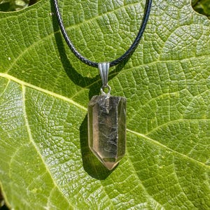 Puede incluir: Un colgante de cristal de cuarzo ahumado cuelga de un collar de cordón negro. El cristal es de color marrón claro y está facetado. El collar se muestra sobre una gran hoja verde.