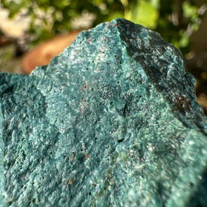 Pode incluir: Um espécime mineral rugoso e texturizado com uma cor verde-azulada vibrante. A superfície é caracterizada por numerosos pequenos orifícios e poros irregulares, com subtis brilhos cristalinos e manchas ocasionais de cor castanho-escura ou avermelhada.