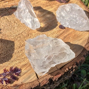 May include: Three clear quartz crystals of varying sizes and shapes, resting on a wooden surface. The crystals are translucent and have a natural, rough texture.