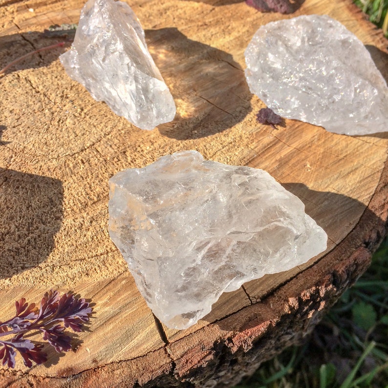 May include: Three clear quartz crystals of varying sizes and shapes, resting on a wooden surface. The crystals are translucent and have a natural, rough texture.
