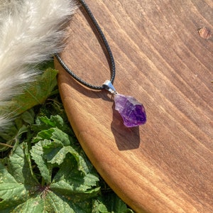 Può includere: Una collana con pendente di cristallo di ametista grezza. Il cristallo viola è sospeso a un cordino nero. Il pendente è incastonato in un metallo color argento. La collana è esposta su una superficie di legno con fogliame verde.