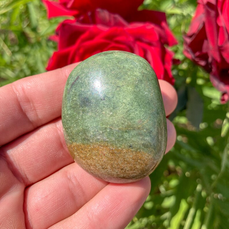 May include: A polished, oval-shaped stone with a smooth surface. The stone features a blend of green and brown hues, with a gradient effect. The stone is held in a hand, with red roses in the background.