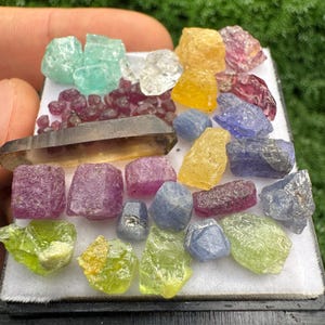 May include: A collection of raw, multi-coloured gemstones arranged on a white display tray. The assortment includes various shapes and sizes of rubies, sapphires, peridot, and a single elongated smoky quartz crystal in shades of red, blue, yellow, and green.