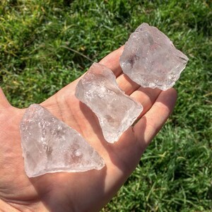 May include: Three clear quartz crystals of varying sizes and shapes, held in a hand against a green grassy background.