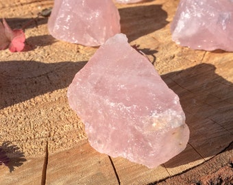 May include: A large, raw, pink rose quartz crystal on a wooden surface. The crystal has a smooth, polished surface and is translucent.