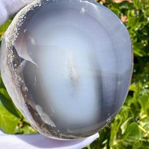 Puede incluir: Una piedra de ágata gris translúcida y pulida con una superficie lisa y redondeada. El interior muestra un centro blanco suave y nublado que se transforma en tonos grises más profundos hacia los bordes, con bandas y texturas naturales.