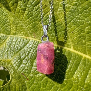 Könnte beinhalten: Ein Anhänger aus rohem, rosafarbenem Rubin hängt an einer filigranen Silberkette. Der Edelstein hat eine rechteckige Form und eine raue, natürliche Textur. Die Halskette liegt auf einem großen, grünen Blatt, mit Schatten.