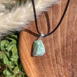 Pode incluir: Um pingente de pedra bruta de cor verde-clara e formato irregular, suspenso por um cordão preto simples. O pingente possui uma argola de metal prateado que conecta a pedra ao cordão, exibindo uma textura natural e não polida.