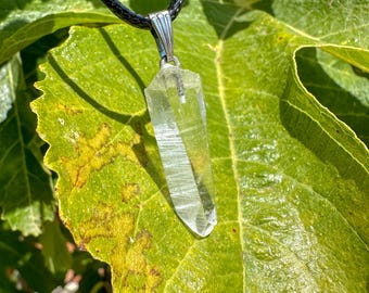 Puede incluir: Un colgante de cristal de cuarzo transparente en un cordón de cuero negro. El cristal está facetado y tiene una punta puntiaguda. El colgante está colgado de una argolla de plata.