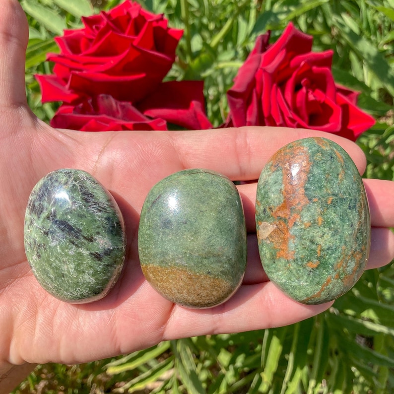 May include: Three polished, oval-shaped green stones with varying patterns and colors are displayed. The stones are held in a hand, with red roses in the background. The stones have a smooth, glossy finish.