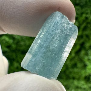 Puede incluir: Un cristal de aguamarina en bruto, translúcido y de color azul claro, con forma prismática hexagonal. La piedra preciosa presenta estrías verticales naturales a lo largo de su cuerpo alargado, con una coloración azul hielo clara y un borde superior ligeramente irregular.