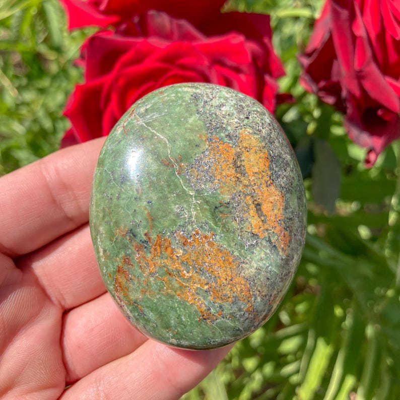 May include: Oval-shaped green stone with orange and gray markings. The polished surface of the stone reflects light, highlighting its natural patterns. The stone is held in a hand, with red roses in the background.