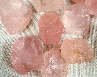 May include: A collection of raw rose quartz crystals in varying shades of pink. The rough, natural stones are displayed on a white, textured surface, showcasing their unique shapes and subtle color variations.