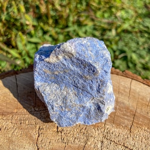 Puede incluir: Un cristal de lapislázuli áspero, azul grisáceo con vetas blancas. El cristal está sentado sobre un trozo de madera.