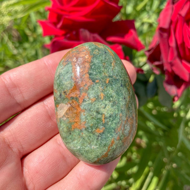 May include: A polished, oval-shaped stone with a mottled green and brown pattern. The stone is held in a hand, with red roses and green foliage in the background. The stone's surface has a natural, earthy appearance.