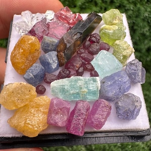 May include: A collection of raw, unpolished gemstones in various shapes and sizes, including yellow, blue, pink, green, and dark brown crystals. The stones are arranged on a white display pad, showcasing natural textures, facets, and colours.