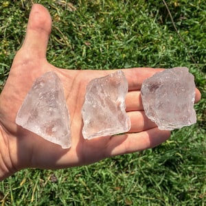 May include: Three clear quartz crystals of varying sizes and shapes, displayed on a hand against a green grassy background.