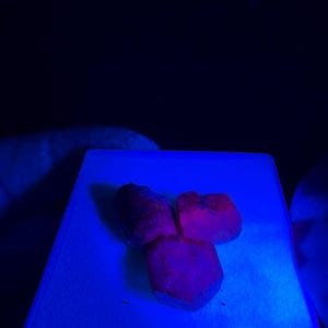 Könnte beinhalten: Drei raue, rötlich-violette Edelsteine werden auf einer weißen Oberfläche unter blauem Licht gezeigt. Die Steine haben unregelmäßige Formen und ein strukturiertes Aussehen, mit einem dunklen Hintergrund.