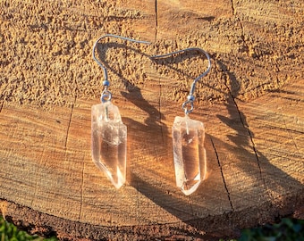 May include: A pair of clear quartz crystal earrings with silver-colored hooks. The crystals are irregularly shaped and have a translucent appearance. The earrings are displayed on a textured, brown wooden surface.