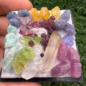 May include: A collection of assorted raw gemstones arranged on a white display pad. The assortment includes blue, yellow, pink, green, and clear crystals of varying shapes and sizes, featuring a prominent elongated clear quartz point in the centre.
