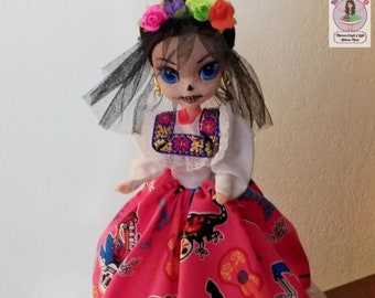 May include: A Day of the Dead doll wearing a white dress with a pink skirt and a black veil. The skirt has a colorful pattern of skeletons, flowers, and musical instruments. The doll has a sugar skull face paint and a flower crown.