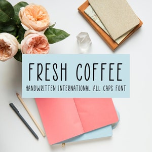May include: A flat lay image with a light blue background featuring a handwritten font design with the text "FRESH COFFEE" in black. The text is accompanied by the phrase "HANDWRITTEN INTERNATIONAL ALL CAPS FONT". The image also includes a bouquet of pink roses, a crystal, a notebook, and two pens.
