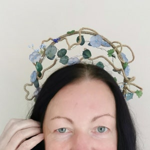 May include: A floral crown with blue and green accents. The crown is made of woven material and features delicate blue flowers and green leaves. The crown is worn on the head.