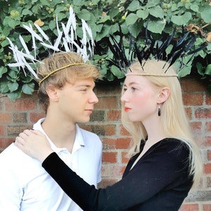 May include: Two people wearing feather crowns. One crown has white feathers, the other has black feathers. The crowns are on a gold and beige band. The background is a brick wall and green foliage.