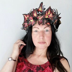 Può includere: Una donna con lunghi capelli neri indossa una corona fatta di foglie rosse, gialle e verdi. Indossa un vestito rosso con un motivo floreale.