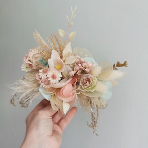 May include: A bouquet of dried flowers in shades of pink, beige, and white. The bouquet includes dried roses, daisies, and other wildflowers. The bouquet is arranged in a hand.