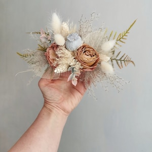 May include: A dried flower arrangement with a blush pink rose, a gray poppy pod, and white pampas grass. The arrangement is held in a hand.