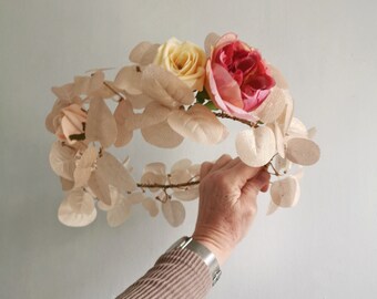Op de afbeelding: Een bloemenkrans gemaakt van beige bladeren en roze en gele bloemen. De krans wordt vastgehouden door een hand met een zilveren horloge.