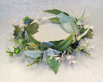 May include: A green and blue floral crown made of artificial leaves and flowers. The crown is made of a white wire base and is decorated with green leaves, blue eucalyptus leaves, and small white flowers.