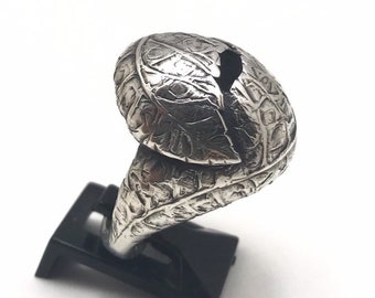 May include: A silver ring with a leaf design. The ring features a detailed texture and a split in the leaf-shaped top. The ring is displayed on a black stand against a white background. The ring's design is intricate and eye-catching.