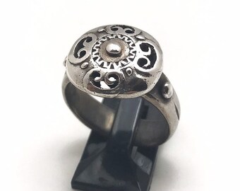 May include: A silver-toned ring with an ornate, domed top featuring a central raised circle and intricate cut-out patterns. The ring is displayed on a black stand against a white background. The ring's design suggests a vintage or antique style.