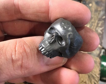 May include: A small, dark gray skull sculpture with detailed features, including eye sockets, teeth, and a star-shaped design on the forehead. The skull is held in a hand, showcasing the intricate craftsmanship of the miniature artwork.