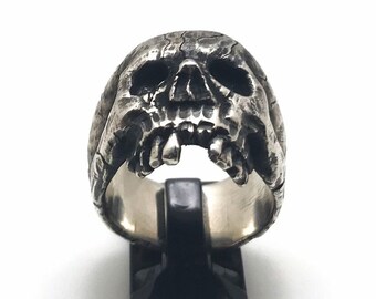 May include: A silver-colored skull ring with a textured surface and open mouth, displaying teeth. The ring is displayed on a black stand against a white background. The ring's design is detailed and gothic.