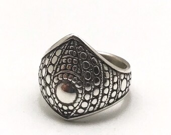 May include: A silver-colored ring with a textured, geometric design. The ring features a central, raised circular element and a pattern of small, interconnected circles. The ring's design is symmetrical and detailed, with a polished finish.