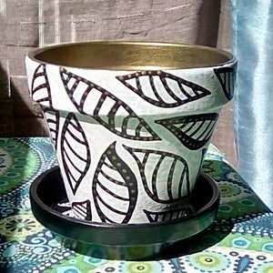 May include: A white ceramic planter with a black and gold leaf design. The planter has a black plastic saucer.