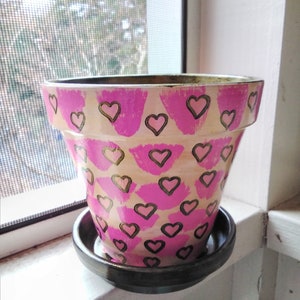 May include: A pink and gold heart patterned terracotta flower pot with a matching saucer.