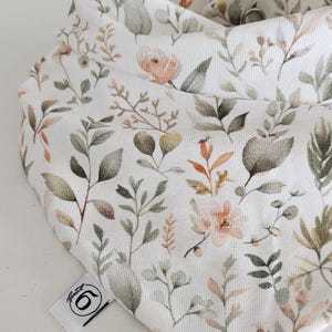 Könnte beinhalten: Ein weißes, dreieckiges Lätzchen mit einem botanischen Druck aus gedämpften grünen Blättern und zarten pfirsichfarbenen Blüten. Der Stoff hat eine sichtbare Textur und ein kleines weißes Etikett an der Seite mit einem schwarzen Logo und der Zahl 6.