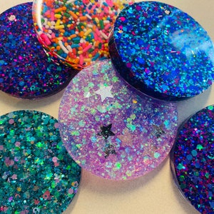 May include: Six round, flat, resin coasters with glitter embedded in the resin. The coasters are in various colors, including purple, blue, teal, and pink. Some of the coasters have a star design.