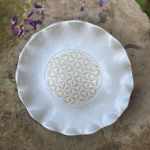 May include: A round, silver-colored dish with a ruffled, wavy edge. The center of the dish features a gold-colored geometric flower of life pattern consisting of multiple overlapping circles arranged in a symmetrical, hexagonal grid design.