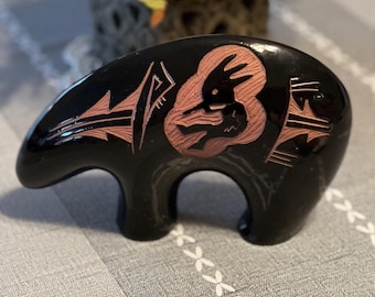May include: Black ceramic bear figurine with a carved design of a Native American figure in a pink color. The bear is standing on a gray surface.