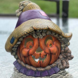 May include: A ceramic Halloween decoration shaped like a smiling jack-o'-lantern wearing a straw hat with a purple band. The pumpkin is orange with a carved face, and the hat has a purple brim and a blue patch.